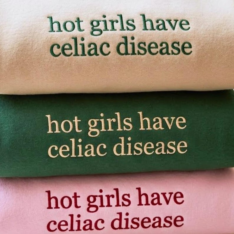 May include: A stack of three folded sweatshirts in beige, dark green, and light pink. Each sweatshirt features the embroidered text "hot girls have celiac disease" in a contrasting color: dark green on beige, beige on green, and dark red on pink.
