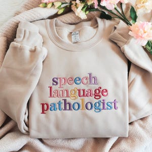 Puede incluir: Una sudadera beige con las palabras "speech language pathologist" bordadas en colores arcoíris.