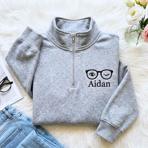 May include: A light gray quarter-zip sweatshirt featuring a black embroidered graphic of eyeglasses with one open eye and one winking eye, positioned above the name Aidan in a black serif font on the left chest area.