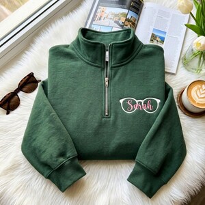 May include: A dark green quarter-zip sweatshirt featuring a silver metal zip and a white graphic of spectacles with the name Sarah written in pink cursive script on the left chest area. The garment has long sleeves with ribbed cuffs.