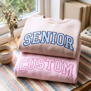 Puede incluir: Dos sudaderas de cuello redondo apiladas una sobre otra. La sudadera superior es de color tostado claro con la palabra SENIOR en letras de bloque de cuadros vichy azules. La inferior es rosa claro con la palabra CUSTOM en cuadros vichy rosas.
