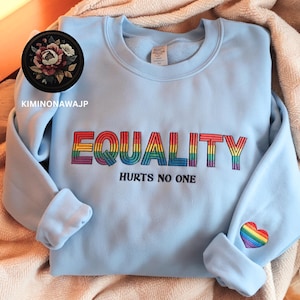 Puede incluir: Sudadera azul claro con las palabras "EQUALITY HURTS NO ONE" bordadas en colores arcoíris. Un corazón arcoíris está bordado en la manga.