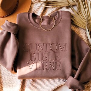 May include: A mauve-colored sweatshirt with the words "CUSTOM BIBLE VERSE" embroidered on the front. A gold chain necklace rests on the neckline. A brown hat and dried palm leaves are in the background.