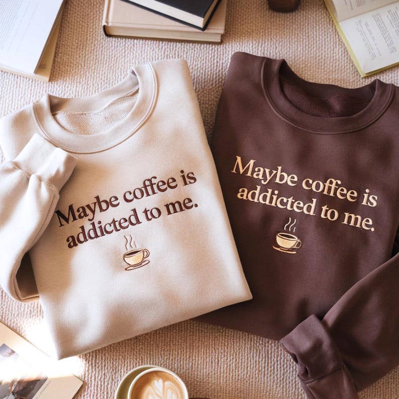May include: Two crewneck sweatshirts, one in cream and one in dark brown, each featuring the text "Maybe coffee is addicted to me." and a small graphic of a steaming coffee cup. The cream sweatshirt has dark brown text, while the brown sweatshirt has light tan text.