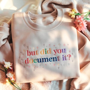 May include: A beige sweatshirt with the embroidered text "but did you document it?" in rainbow colors. The sweatshirt has a crew neck and is decorated with small embroidered starbursts. The garment is folded on a soft, textured surface.