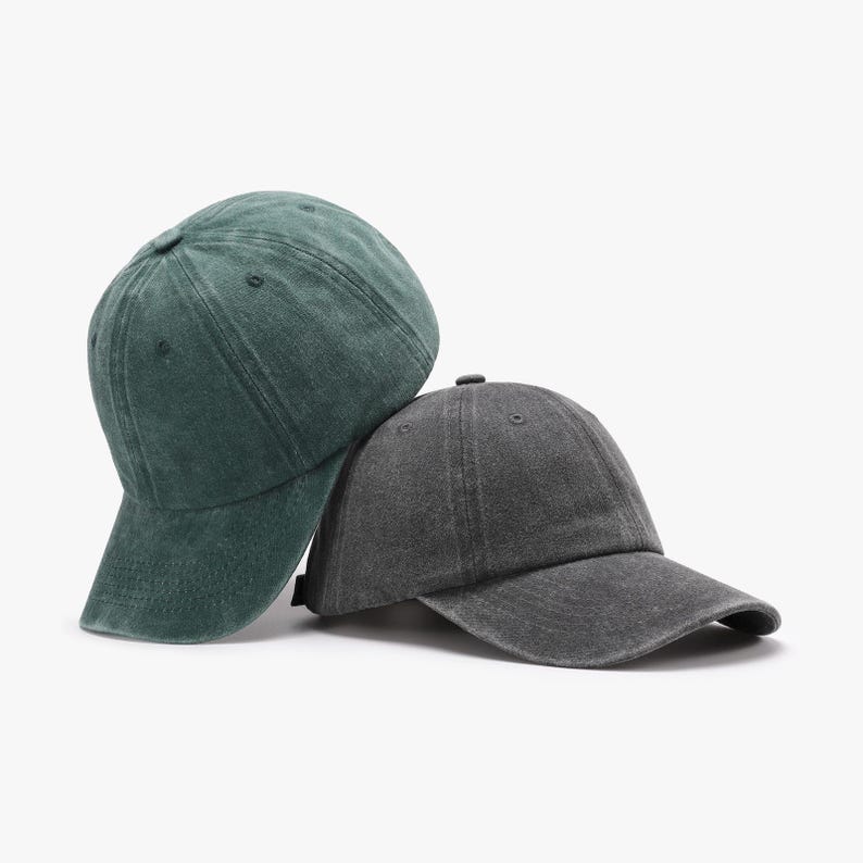 May include: Two baseball caps, one forest green and one charcoal gray, are displayed. The caps have a classic six-panel design with a curved brim. The fabric appears to be a washed cotton or similar material, giving them a slightly worn look.