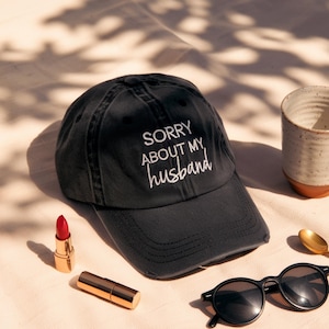 May include: A black baseball cap with the text "SORRY ABOUT MY husband" embroidered in white. The cap is displayed with a red lipstick, a gold lipstick tube, black sunglasses, a gold spoon, and a ceramic mug.