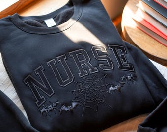 May include: Black sweatshirt with the word "NURSE" in a gothic font. The design includes a spider web, bats, and spiders, creating a Halloween theme. The sweatshirt is made of a soft material.