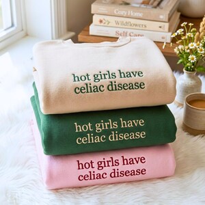 May include: A stack of three crewneck sweatshirts in beige, dark green, and light pink. Each sweatshirt features the embroidered text "hot girls have celiac disease" in a contrasting color, with dark green, cream, and dark red lettering respectively.