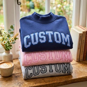 May include: A stack of three crewneck sweatshirts in navy blue, light pink, and heather grey. Each sweatshirt features the word CUSTOM in large, block-style letters with a gingham pattern fill, stitched across the centre of the chest area.