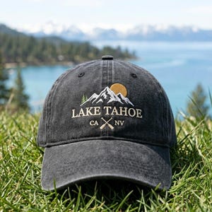 May include: A charcoal grey baseball cap featuring an embroidered design of white mountain peaks, a yellow sun, and green pine trees. The text reads LAKE TAHOE CA NV with two crossed oars below the lettering, all centered on the front panel.