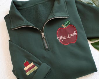 May include: A dark green pullover sweatshirt with a quarter-zip neckline. An embroidered red apple with the text "Mrs Leach" is on the front. The left sleeve cuff features an embroidered stack of books and an apple.