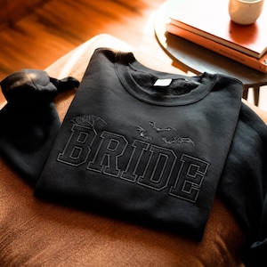 May include: A black crewneck sweatshirt featuring the word BRIDE embroidered in a bold, block-style font across the chest. Above the text, there are three small, detailed embroidered bat designs. The garment has long sleeves and a ribbed neckline.