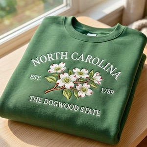 May include: A forest green crewneck sweatshirt featuring white embroidered text that reads NORTH CAROLINA EST. 1789 THE DOGWOOD STATE. Centered is an embroidered illustration of white dogwood flowers with green leaves and brown stems.