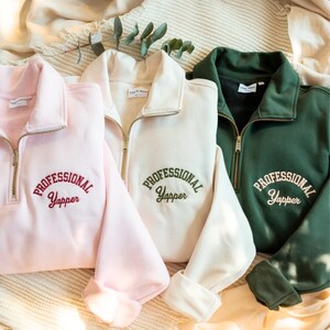 May include: Three quarter-zip sweatshirts in pink, cream, and green. Each sweatshirt has the embroidered text "PROFESSIONAL Yapper" across the chest. The sweatshirts have a collared neckline and long sleeves. The background is a neutral colour.