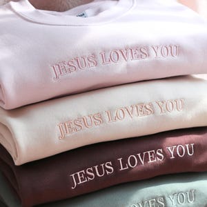 May include: A stack of pastel-colored sweatshirts. Each sweatshirt is embroidered with the phrase "JESUS LOVES YOU" in a matching thread color. The colors include light pink, cream, burgundy, and a muted green. The sweatshirts are neatly folded.