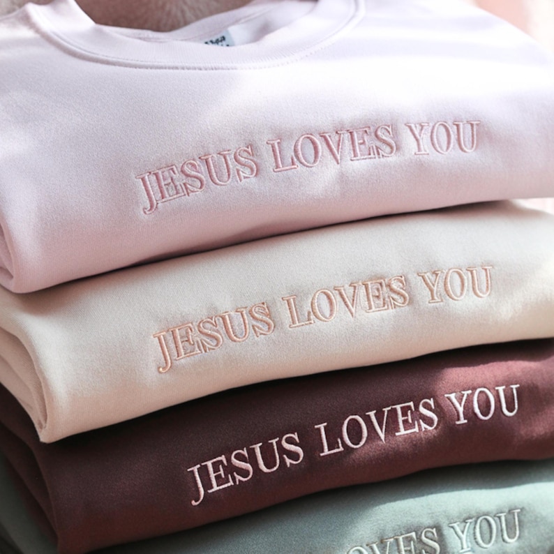 May include: A stack of pastel-colored sweatshirts. Each sweatshirt is embroidered with the phrase "JESUS LOVES YOU" in a matching thread color. The colors include light pink, cream, burgundy, and a muted green. The sweatshirts are neatly folded.