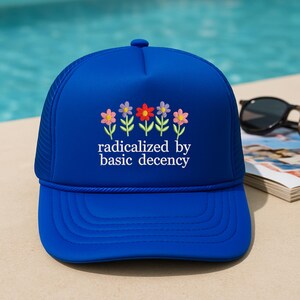 Puede incluir: Gorra de camionero azul real con visera curva y parte trasera de malla. El frente presenta un diseño bordado de flores coloridas y el texto "radicalized by basic decency" en blanco. Un accesorio de moda para uso diario.
