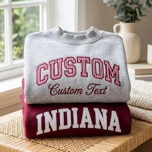 May include: A light gray crewneck sweatshirt with the word CUSTOM in red and white gingham block letters and the text Custom Text below it, stacked on a maroon sweatshirt with the word INDIANA in large white block letters.
