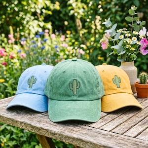 May include: Three baseball caps in light blue, sage green, and mustard yellow, each featuring a small embroidered green cactus icon on the front panel. The caps have a structured crown, a curved brim, and a washed fabric texture.