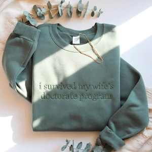 May include: Sage green sweatshirt with the phrase "i survived my wife's doctorate program" embroidered on the front. A gold chain necklace rests on the sweatshirt. The sweatshirt is folded on a white surface.