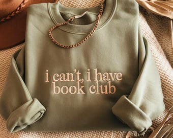 May include: Sage green sweatshirt with the embroidered text "i can't, i have book club." A gold chain necklace is draped across the sweatshirt. The sweatshirt is folded on a textured, cream-colored surface.