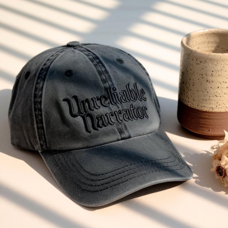 May include: A grey denim baseball cap featuring the embroidered phrase "Unreliable Narrator". The cap has a curved peak and a button on top. A ceramic mug is visible in the background.