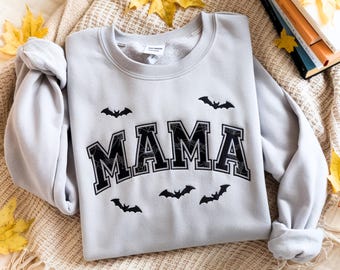 May include: A light gray sweatshirt with the word "MAMA" in black, outlined with white, and filled with a spiderweb design. Small black bat silhouettes are scattered around the word. The sweatshirt is folded on a beige blanket.