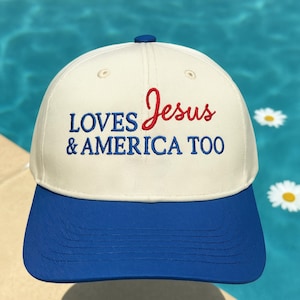 May include: A baseball cap with a white crown and a blue brim. The cap features the embroidered text "LOVES Jesus & AMERICA TOO" in red and blue thread. The cap has a blue button on top.