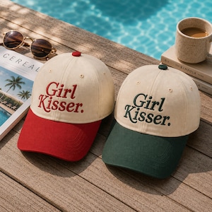 May include: Two baseball caps with cream-colored crowns and embroidered text that reads Girl Kisser. One cap features a red brim and red lettering, while the other cap features a dark green brim and dark green lettering. Both caps have a button top.