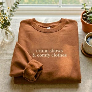 May include: A brown sweatshirt with the text "crime shows & comfy clothes" in a white font. The sweatshirt is folded, with a sleeve cuff visible. The background includes a window, plants, and a cup of coffee.