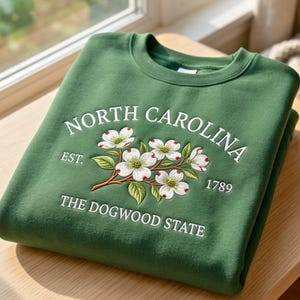 May include: A forest green crewneck sweatshirt featuring white embroidered text that reads NORTH CAROLINA EST. 1789 THE DOGWOOD STATE. Centered is an embroidered illustration of white dogwood flowers with green leaves and brown stems.