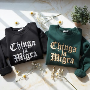 May include: Two crewneck sweatshirts, one black and one dark green, with the embroidered text "Chinga la Migra" in a gothic font. The black sweatshirt has white embroidery, while the green one has gold embroidery. Daisies and a small potted plant are in the background.