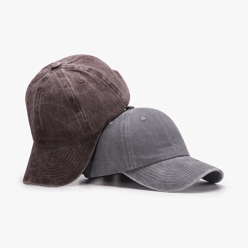 May include: Two baseball caps in a studio setting. One is a faded brown color, and the other is a faded gray. Both caps have a curved brim and a button on top. The caps are made of a soft, worn material.