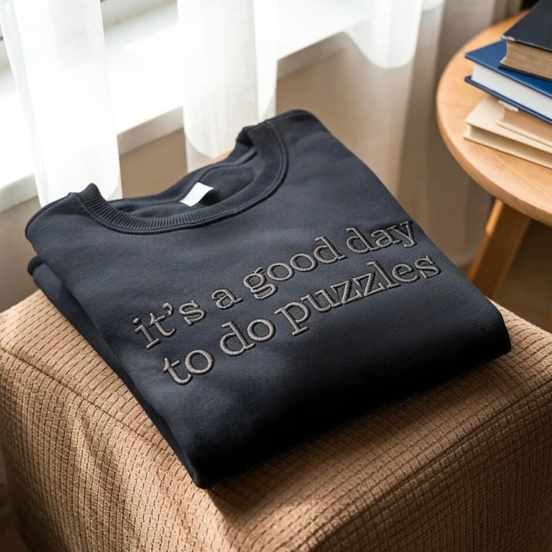 May include: A dark navy blue sweatshirt with the embroidered text "it's a good day to do puzzles". The sweatshirt is folded and resting on a brown, textured surface. The image is well-lit, with a soft focus.