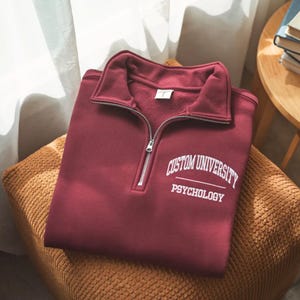 May include: A maroon sweatshirt with a quarter-zip collar and the text "CUSTOM UNIVERSITY PSYCHOLOGY" in white. The sweatshirt is folded and resting on a textured, brown surface.