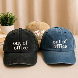 May include: Three baseball caps in different colors: black, blue, and beige. Each cap has the phrase "out of office" embroidered in white on the front.
