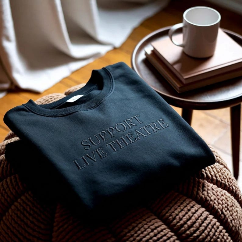 May include: A dark navy blue sweatshirt with the words "SUPPORT LIVE THEATRE" embroidered across the front. The sweatshirt is folded on a woven brown ottoman. A small wooden side table with books and a white mug is in the background.