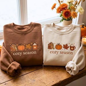 Peut inclure: Deux sweat-shirts à col rond, un marron foncé et un crème, arborant chacun une broderie représentant une pile de livres, une citrouille, une feuille d'érable et une tasse de chocolat chaud avec le texte cozy season centré sous les motifs.