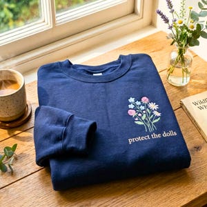 May include: A navy blue sweatshirt with a floral embroidery design and the text "protect the dolls" in a light pink color. The sweatshirt is folded on a wooden surface, with a cup of coffee and a vase of flowers in the background.