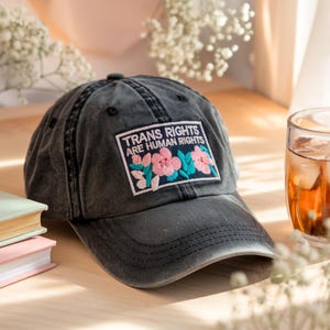 May include: A dark gray baseball cap with a patch that reads "TRANS RIGHTS ARE HUMAN RIGHTS." The patch features pink and teal floral embroidery. The cap is sitting on a wooden surface.