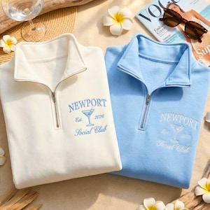 May include: Two quarter-zip sweatshirts, one cream and one light blue, embroidered with "Newport Social Club" and a cocktail glass. The sweatshirts have silver zippers and are displayed with a magazine, sunglasses, and flowers.