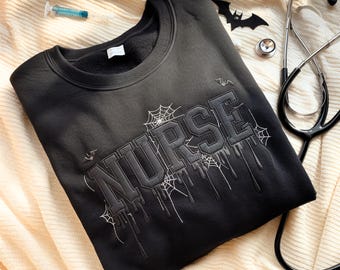 Könnte beinhalten: Schwarzes Sweatshirt mit dem Wort "NURSE" in einem tropfenden Schriftzug, verziert mit Spinnweben und Fledermäusen. Ein Stethoskop und eine Spritze deuten auf ein Halloween-Thema hin.