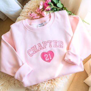 Peut inclure: Un sweat-shirt rose clair avec le mot "CHAPTER" brodé en rose au-dessus d'un cœur rose avec le numéro "21". Le sweat-shirt a un col rond et des manches longues. Des fleurs roses sont en arrière-plan.