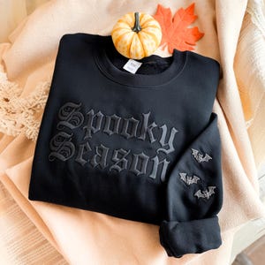 May include: Black sweatshirt with the embroidered text "Spooky Season" in gothic lettering. The sleeve features three embroidered bats. A small pumpkin and an orange leaf are placed on top of the sweatshirt.