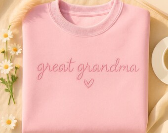 May include: A light pink sweatshirt with the embroidered words "great grandma" in a cursive font, with a small heart below. The sweatshirt has a crew neck and is made of a soft material. The image is taken from an overhead angle.