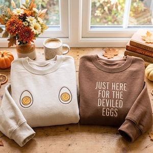 May include: A cream-colored sweatshirt featuring two embroidered halved hard-boiled eggs and a brown sweatshirt with the embroidered text "JUST HERE FOR THE DEVILED EGGS" in a light-colored font, both displayed on a wooden surface.