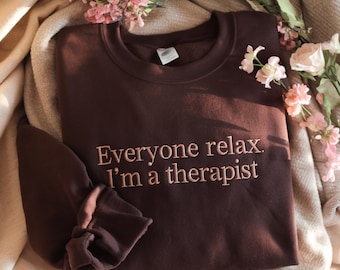 May include: A dark brown crewneck sweatshirt featuring the embroidered text "Everyone relax. I'm a therapist" in a light pink thread. The garment has long sleeves with ribbed cuffs and a ribbed neckline, presented with a white tag at the collar.