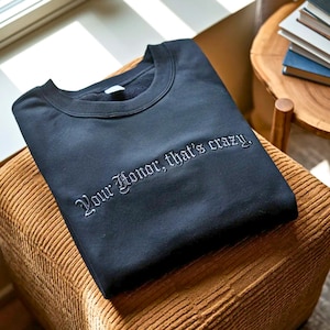 May include: A black sweatshirt with the embroidered text "Your Honor, that's crazy." The crew neck sweatshirt is folded on a brown, textured surface. The sweatshirt is a dark navy blue color.