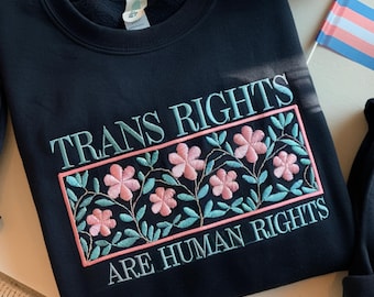 Könnte beinhalten: Ein marineblaues Sweatshirt mit dem gestickten Text "TRANS RIGHTS ARE HUMAN RIGHTS". Das Design zeigt ein rechteckiges Blumenmuster mit rosa Blüten und türkisfarbenen Blättern, umrahmt von einem rosa Rand. Im Hintergrund ist eine Transgender-Pride-Flagge zu sehen.